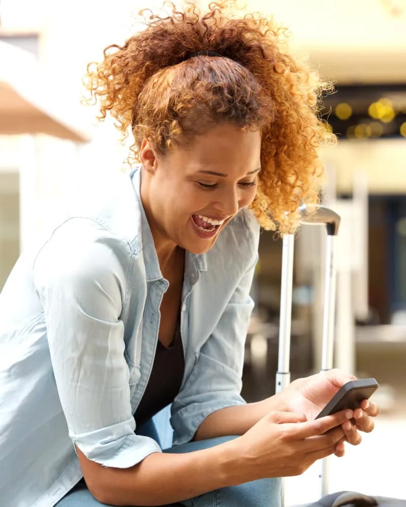 Mulher sentada no aeroporto, rindo enquanto olha o celular, com a mala ao lado