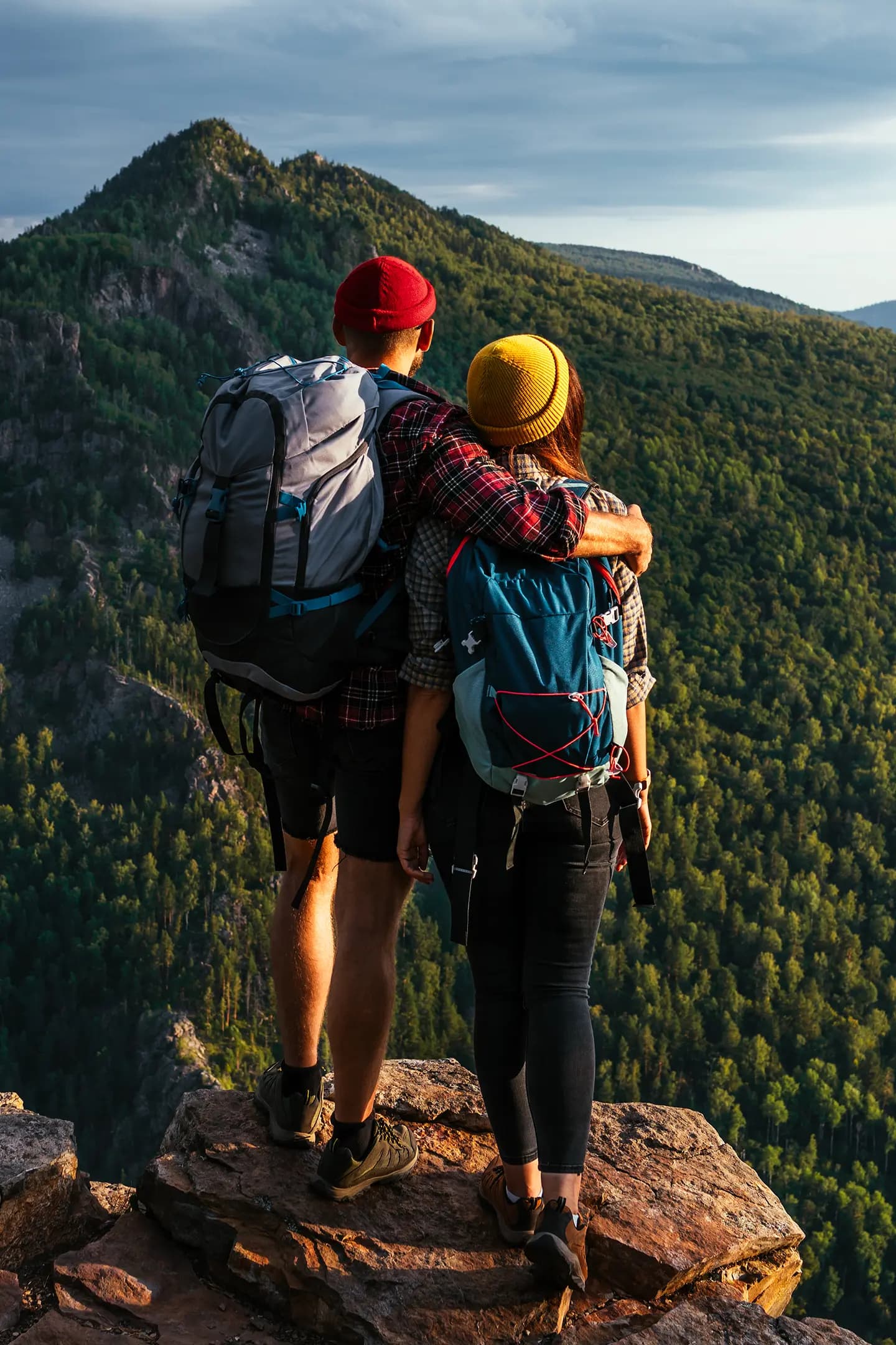 Casal de mochila, de costas e abraçado, na beira de um penhasco olhando uma montanha coberta de mata
